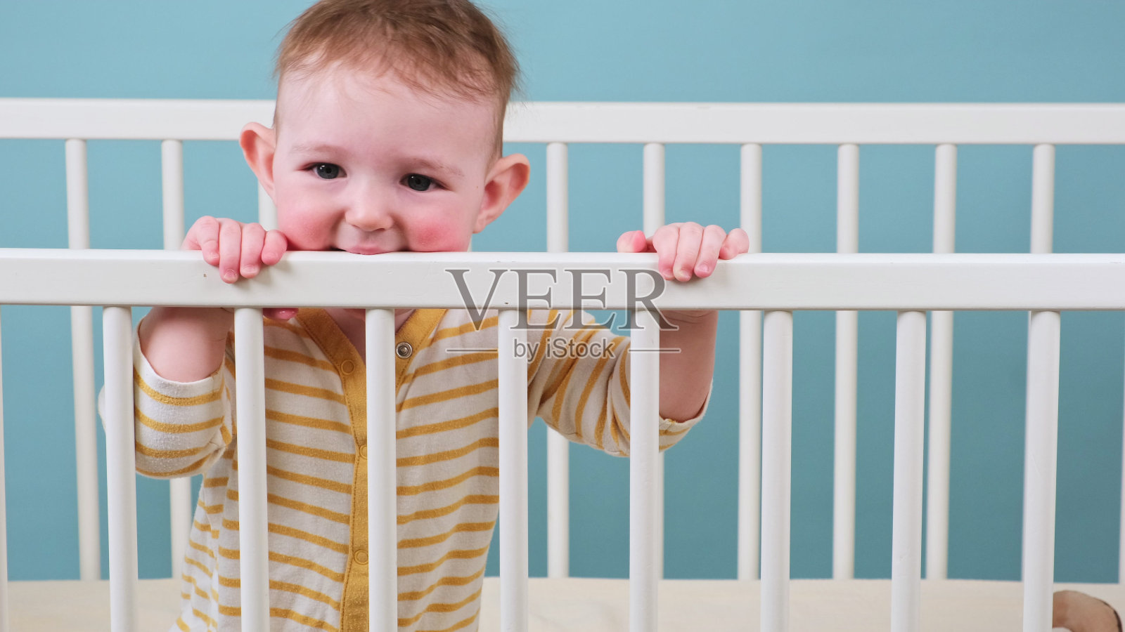 悲伤的蹒跚学步的小男孩在婴儿床上啃着木条，蓝色的画室背景。一个穿着黄色睡衣的疲惫的孩子照片摄影图片