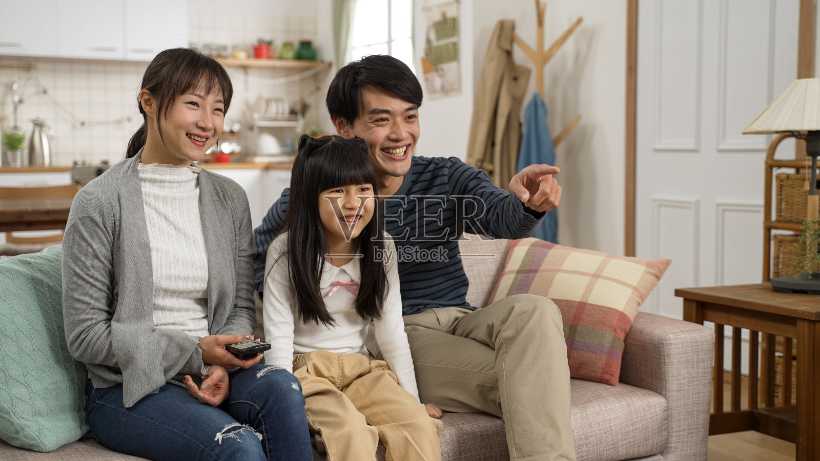 幸福的亚洲三口之家在家里的客厅里一起看电视。可爱的女孩与她的父亲笑，而他是指着电视照片摄影图片