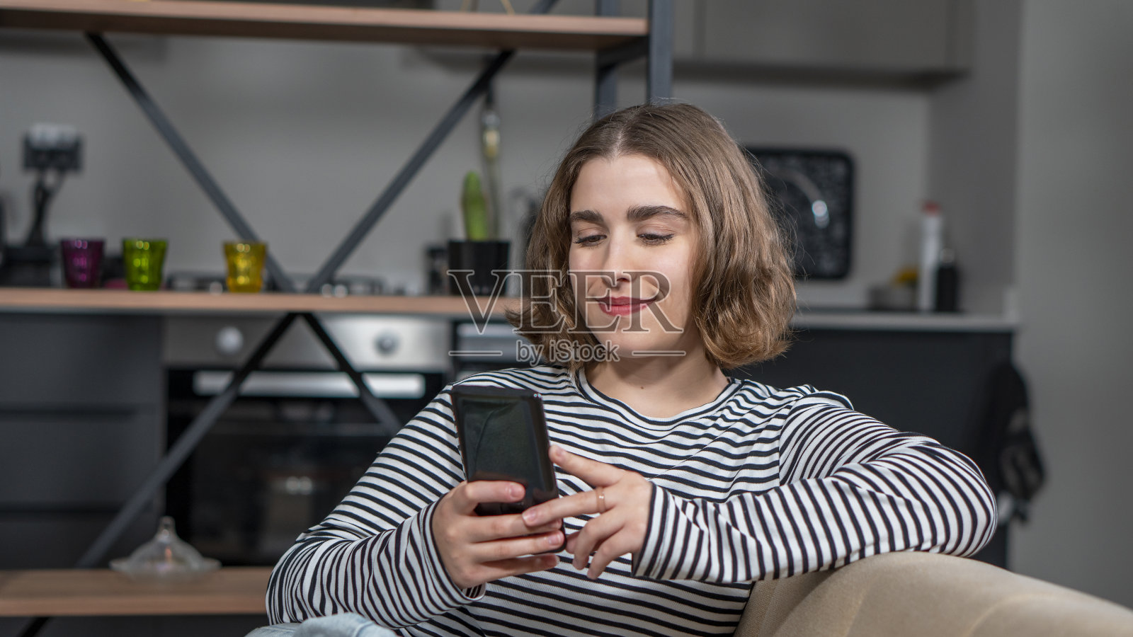 快乐的千禧一代土耳其少女拿着智能手机在家查看社交媒体。微笑的年轻女子用手机app玩游戏，网上购物，点外卖，放松地躺在沙发上。照片摄影图片