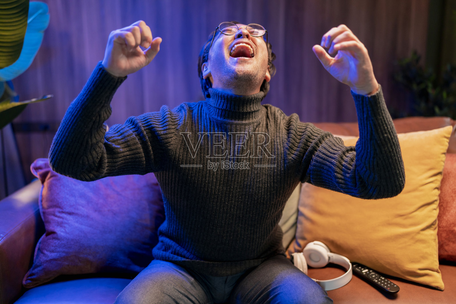 一名亚洲成年男子坐在沙发上看电视上的体育比赛。一名兴奋的男子看着足球比赛，手举起来庆祝进球，在家里高声欢呼照片摄影图片