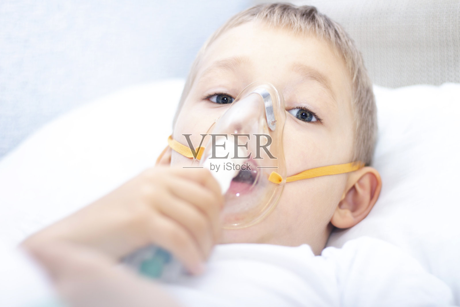 带着吸入器面罩的男孩——患有哮喘的呼吸系统疾病。一个带着呼吸器面罩的男孩躺在床上，呼吸着肾上腺素。保健理念与病儿、冠状病毒、支气管炎、肺炎有关照片摄影图片