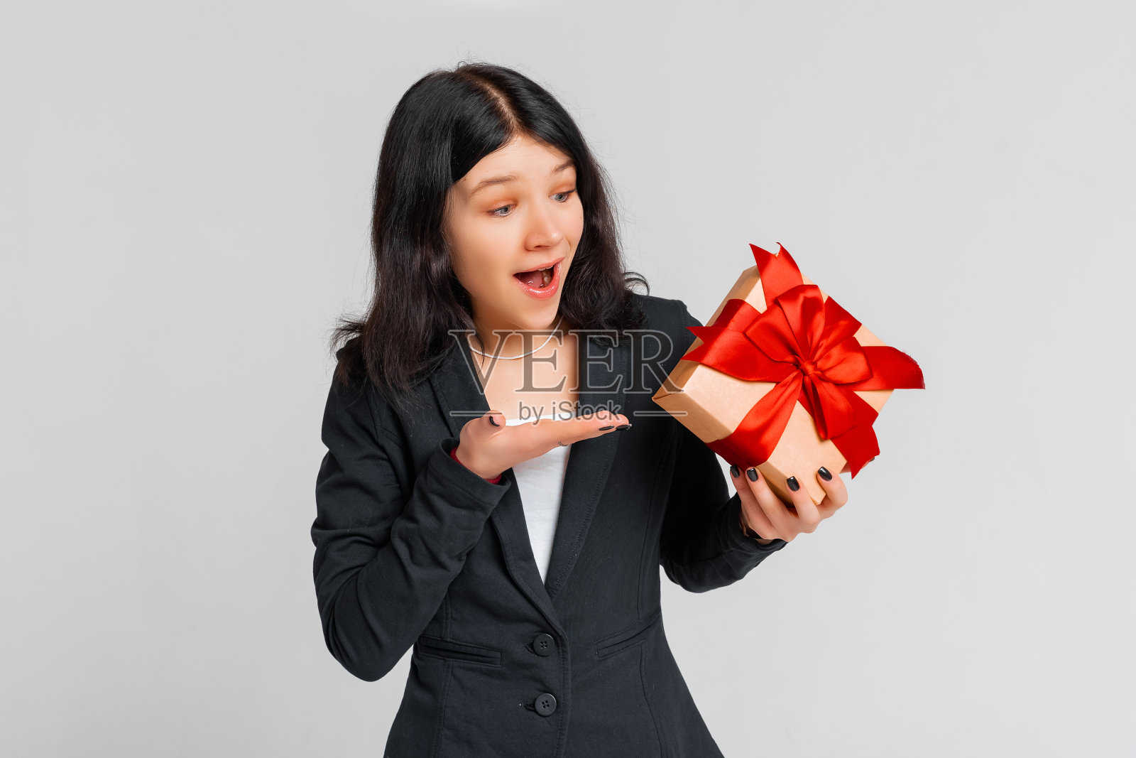 一个穿着黑色外套的黑发女孩好奇地指着一个礼品盒的肖像，焦点，好奇里面是什么，站在灰色的背景上。年轻的情绪化的女人照片摄影图片