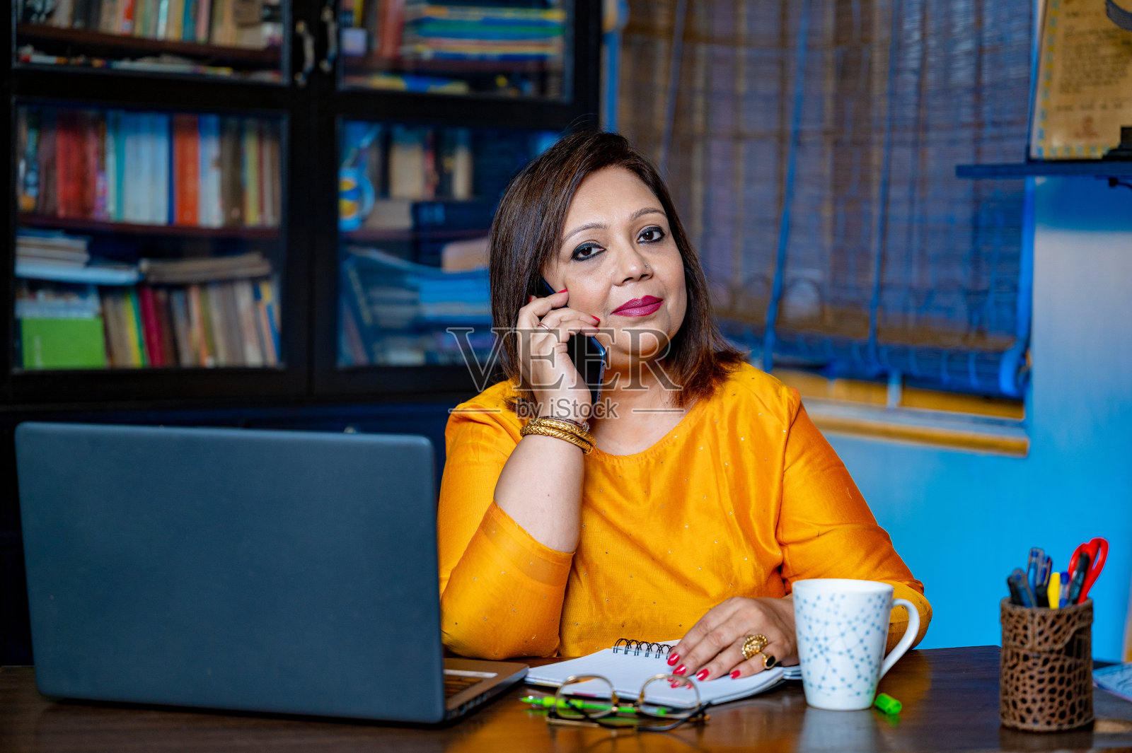 快乐的中年女商人或教师，戴着眼镜坐在家里的办公室，用笔记本电脑打字，用手机。照片摄影图片