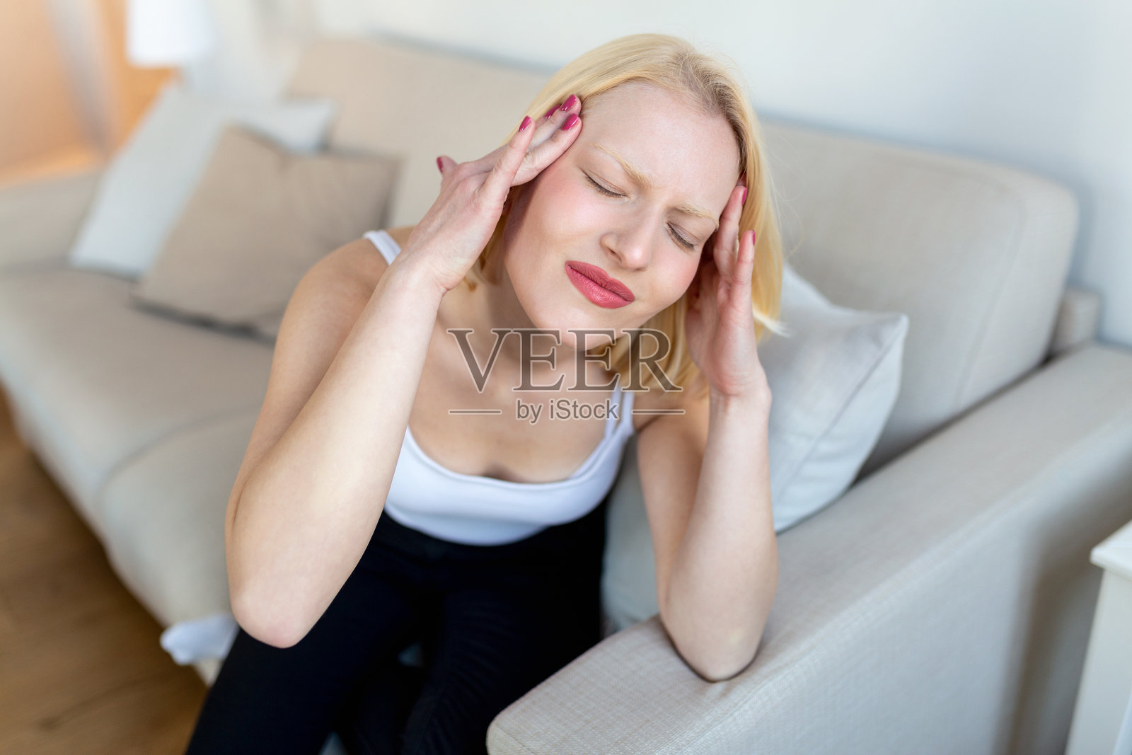 一位年轻的金发女子坐在家里的沙发上，头痛和偏头痛。美丽的女人患有慢性每日头痛。悲伤的女人因为鼻窦疼痛而抱着头照片摄影图片