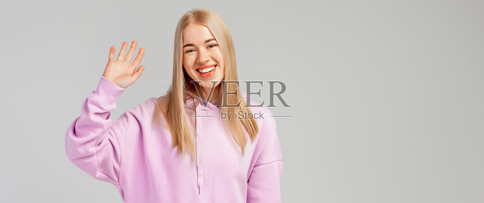 微笑的快乐女孩友好挥手的肖像，说你好，问候，hi手势，站在灰色背景上的紫色连帽衫，复制空间在右侧照片摄影图片