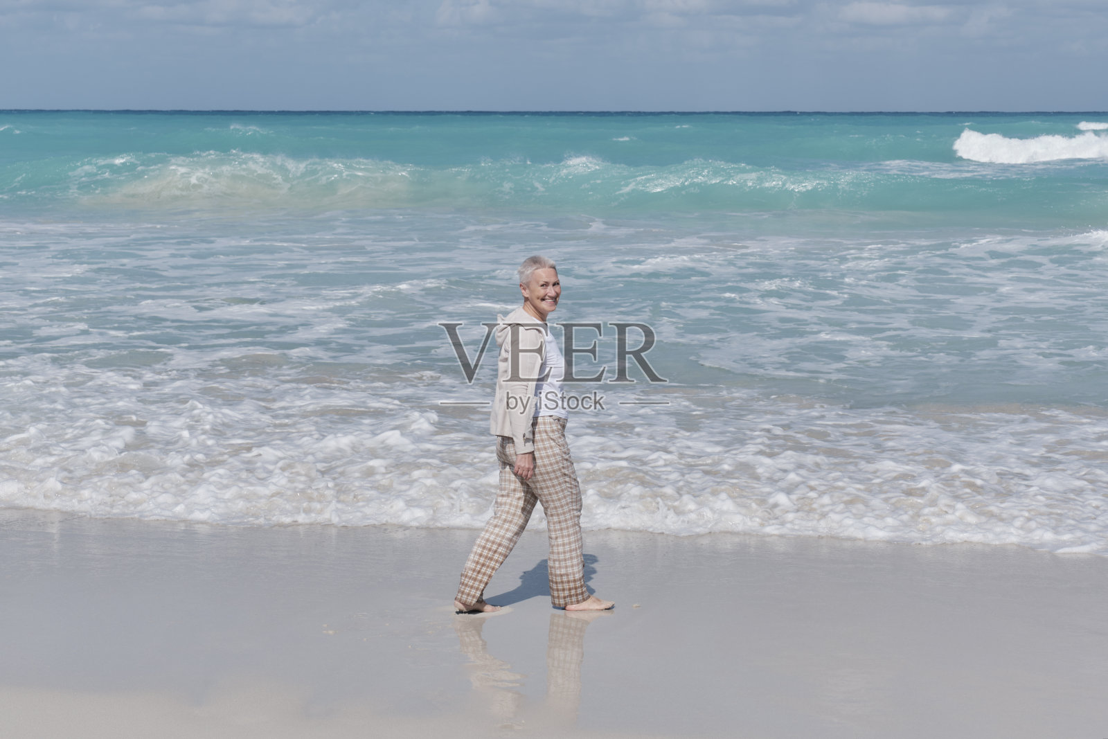 在古巴巴拉德罗大西洋的沙滩上，一名白人老年妇女微笑着走在沙滩上。照片摄影图片