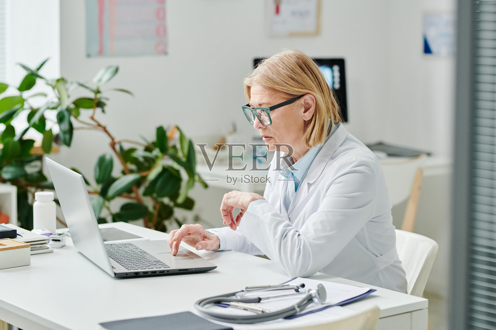 严肃的金发女临床医生穿着白大褂在办公室的笔记本电脑前工作照片摄影图片