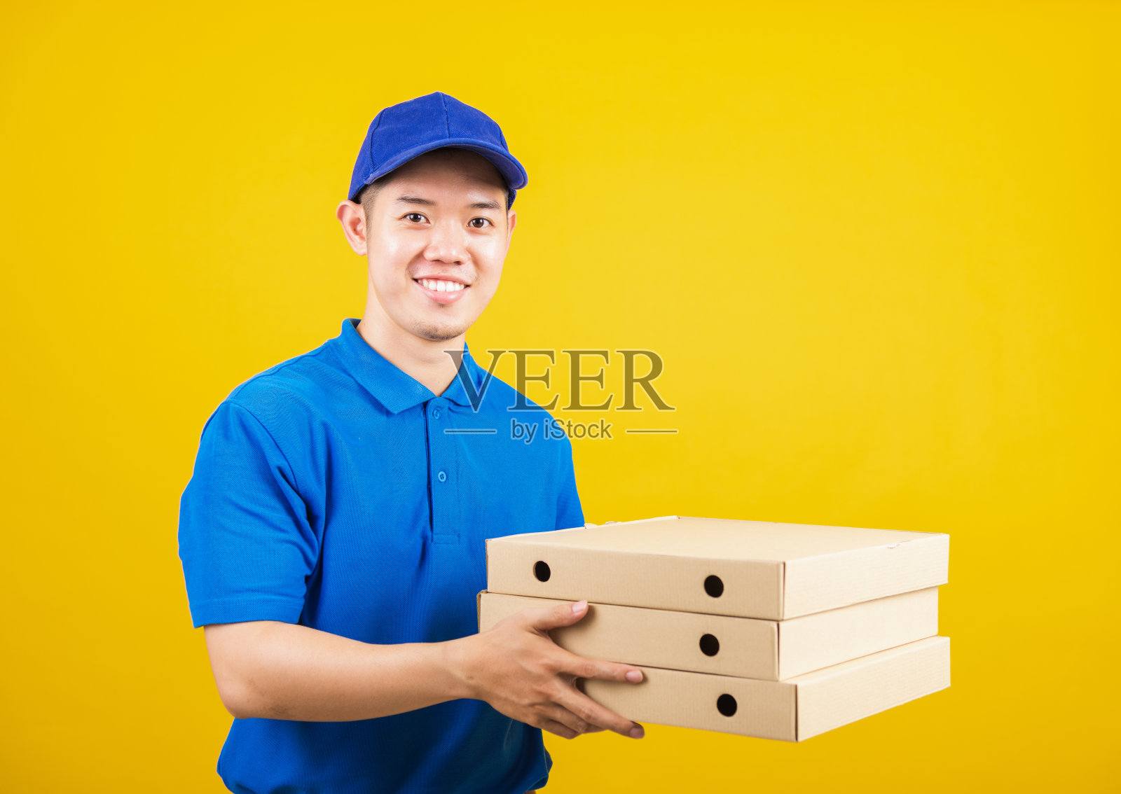 快递员站在那里，他微笑着穿着蓝色t恤，制服戴着帽子，手里拿着送外卖的披萨纸盒照片摄影图片