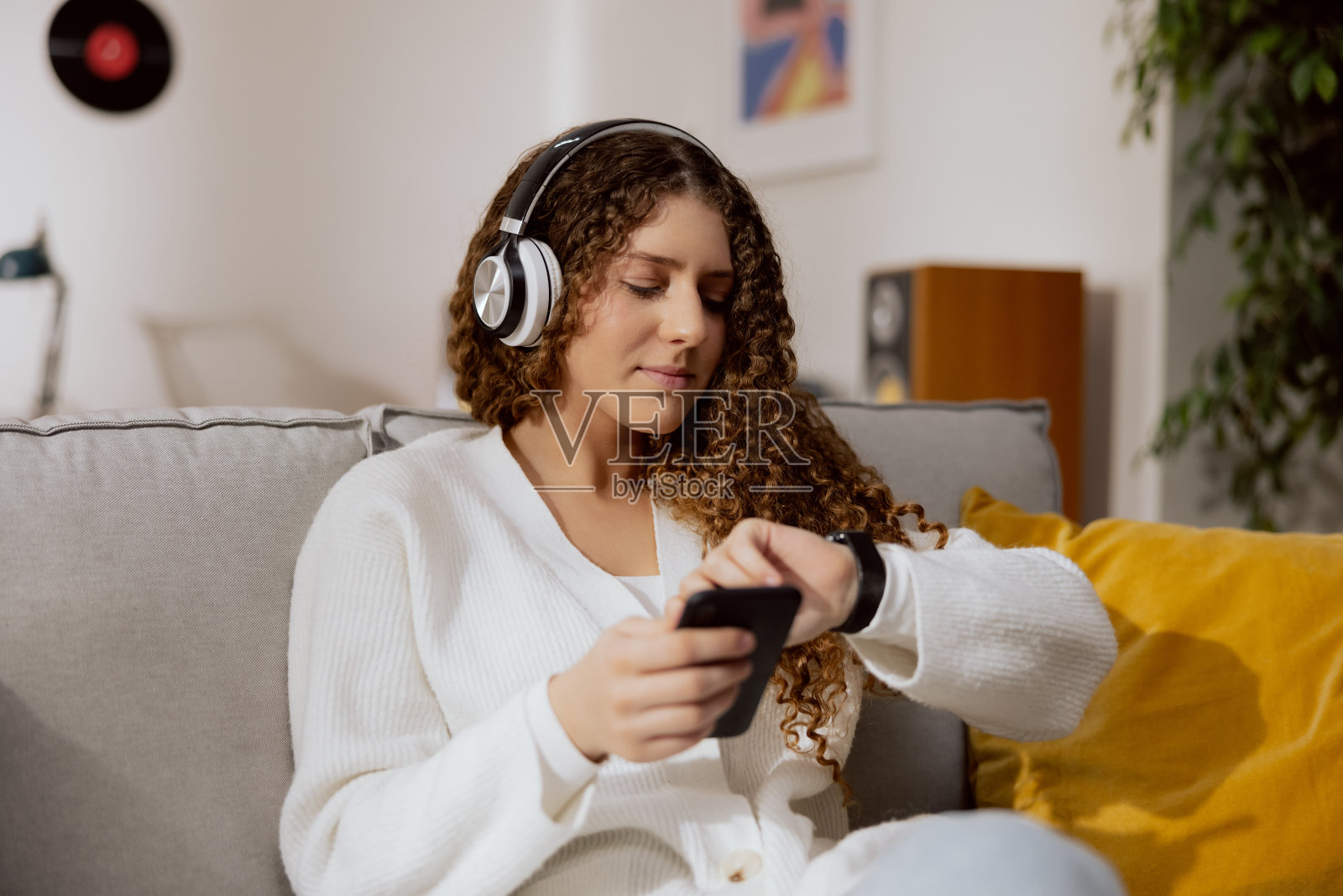 可爱的年轻女孩坐在客厅的灰色舒适的沙发上。右手拿着手机，通过无线耳机听你最喜欢的播客。他看了看左手上的表，以免开会迟到照片摄影图片