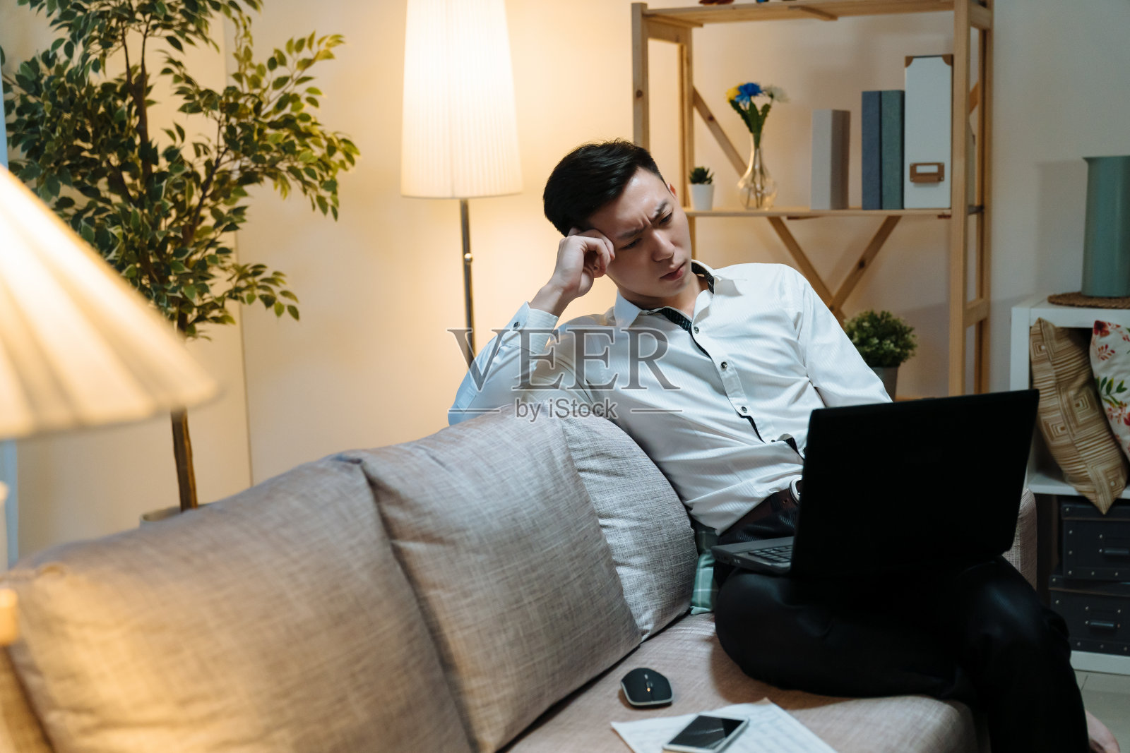 年轻的亚裔韩国商人坐在家里办公室的沙发上，用笔记本电脑工作。业务问题深思熟虑的解决方案概念。深夜忧心忡忡的男员工在沉思。照片摄影图片
