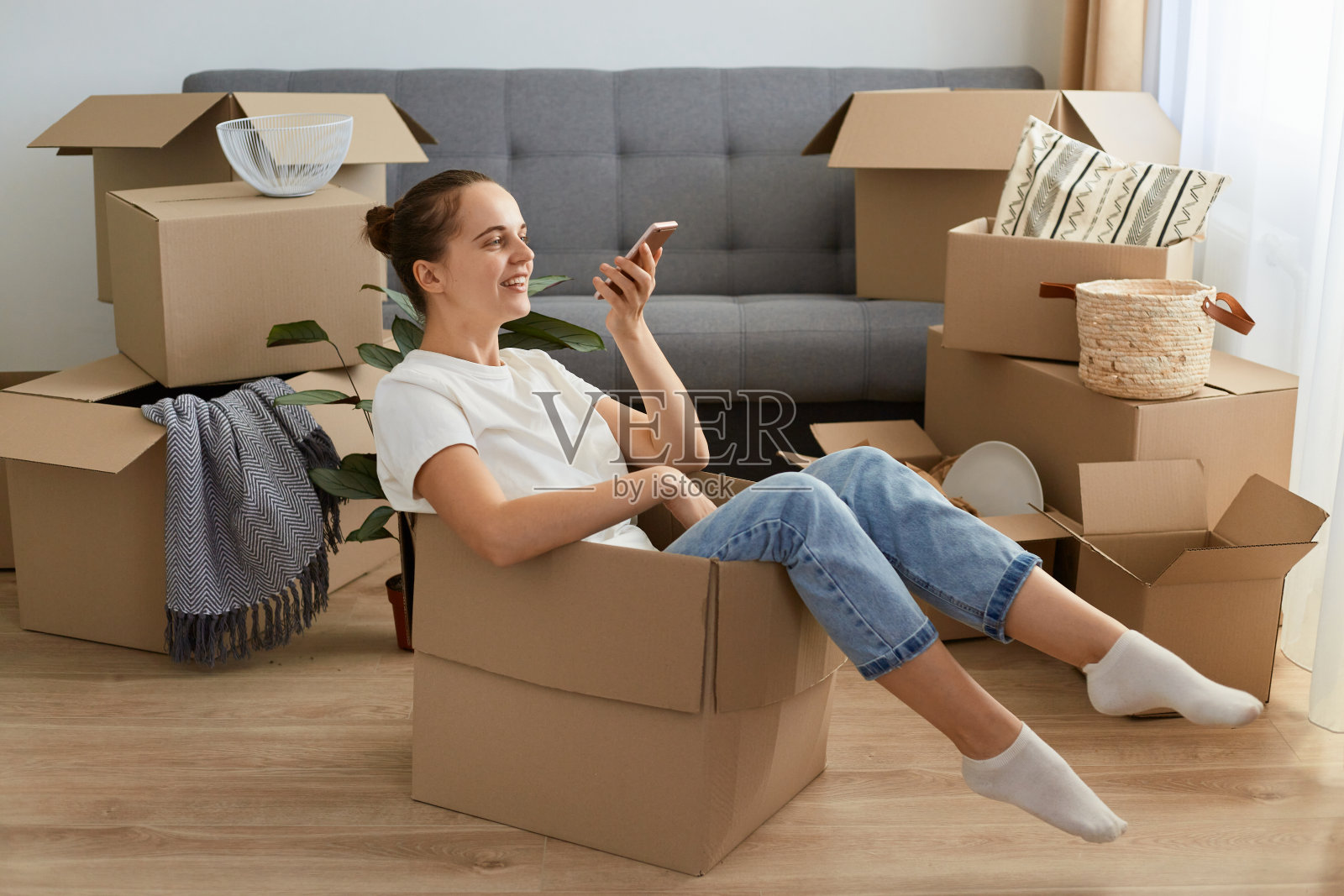 年轻的白人女性穿着白色t恤和牛仔裤，坐在一个纸板箱里，手里拿着智能手机，录制语音信息，讲述她的搬迁。照片摄影图片
