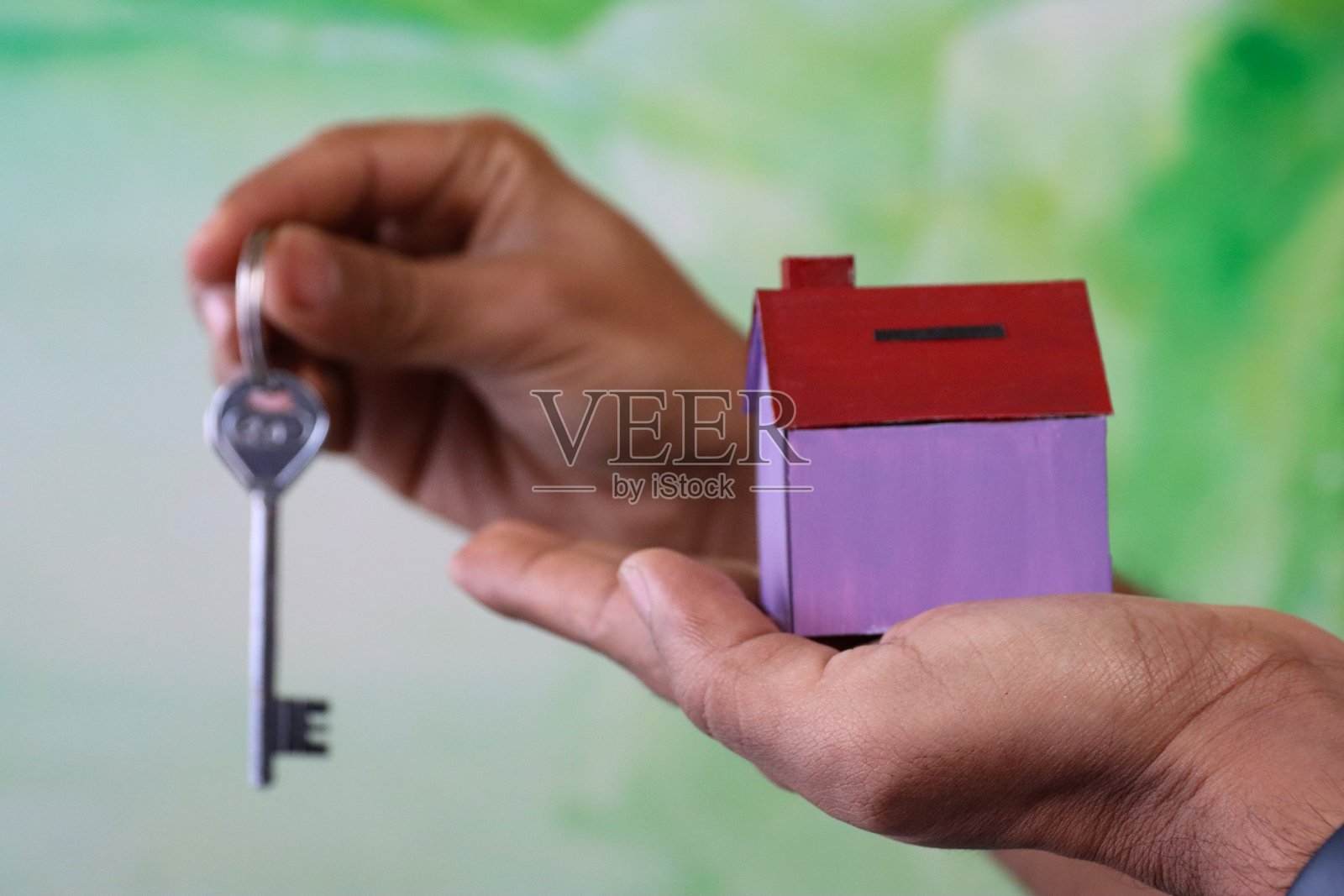 一个陌生的人手持紫色的纸板房子，另一只手挂着钥匙扣，斑驳的绿色背景，房地产和搬家的概念照片摄影图片