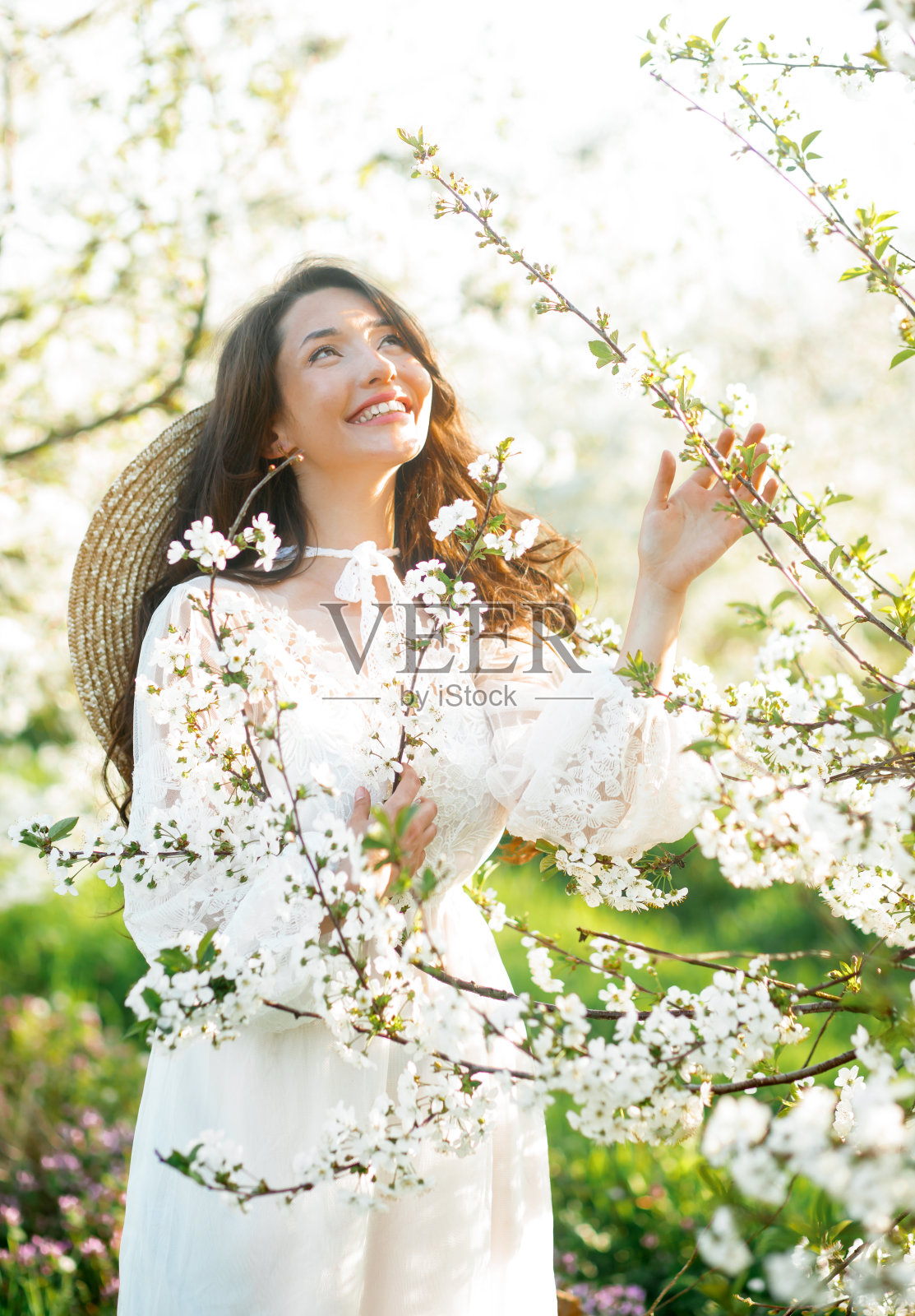 美丽的肖像，一个迷人的年轻女孩在一个开花的花园。头戴草帽、身穿白色连衣裙的卷发女子。照片摄影图片