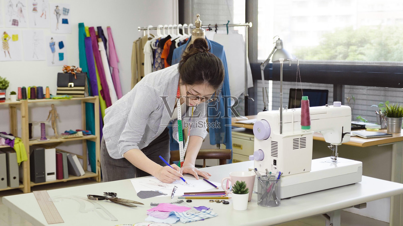 优雅的时装设计师在工作室设计的女人。亚洲女士工人在眼镜持有标记画设计新季节春天收集站在书桌在现代舒适的工作场所。照片摄影图片