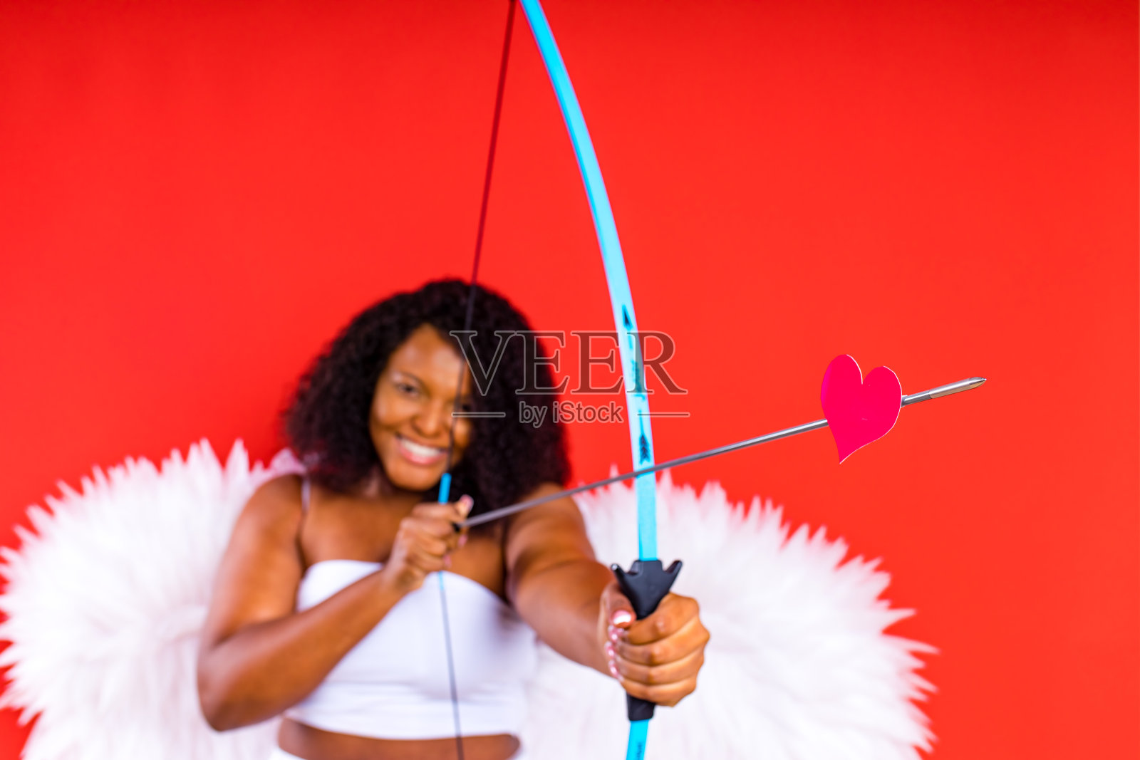 非洲拉丁丘比特女人在白色顶部胸罩拿着弓和箭在红色的工作室2月14日情人节照片摄影图片