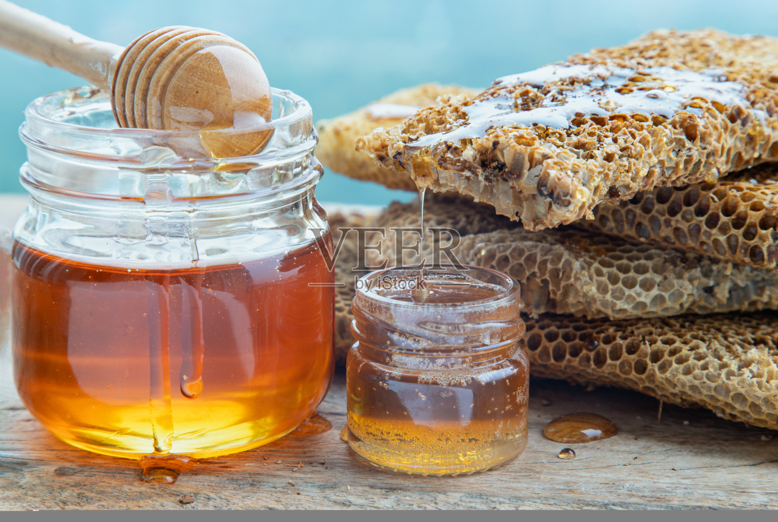 近距离的美味蜂蜜从新鲜的蜂巢滴在玻璃罐与木制蜂蜜勺粘在旧的木制桌子。照片摄影图片