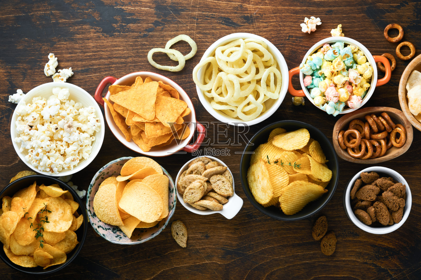 不健康的食物或零食。所有经典的土豆小吃，花生，爆米花，洋葱圈和咸味椒盐脆饼放在碗盘里，在旧的木制背景上。对身材、心脏、皮肤、牙齿都不健康。照片摄影图片