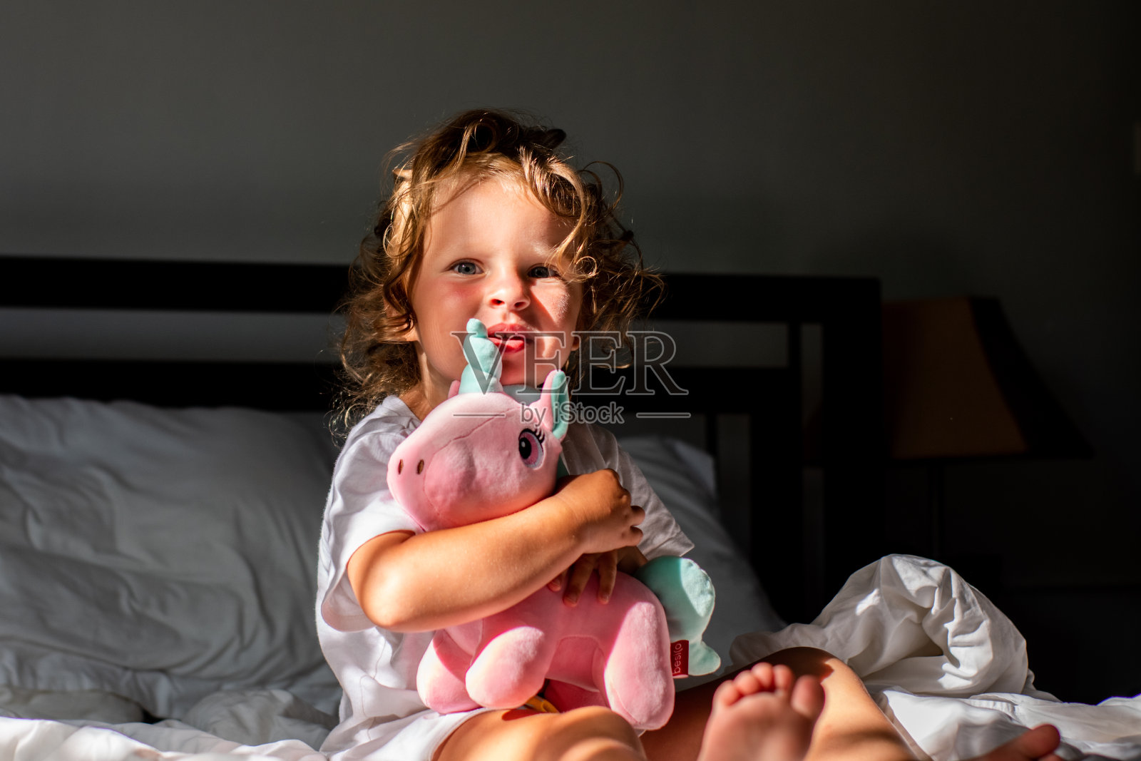 一个卷发的小女孩早上在床上醒来，和一只粉红色的独角兽玩耍。孩子在阳光下醒来。照片摄影图片