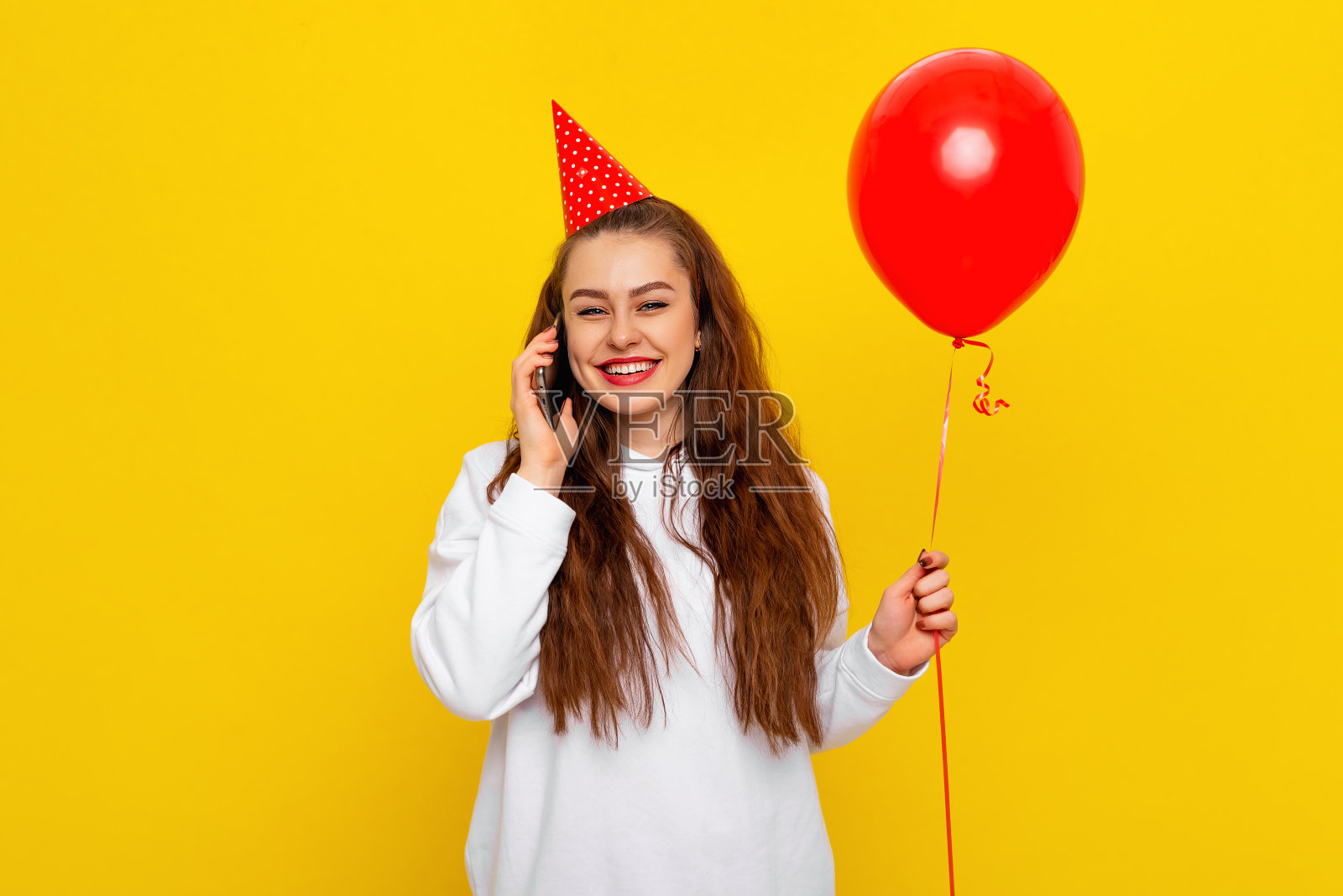 积极的年轻女子正在讲手机，收到朋友们周年庆的祝贺，穿着派对圆锥形和白色休闲风格的毛衣，感觉很高兴，黄色的背景照片摄影图片