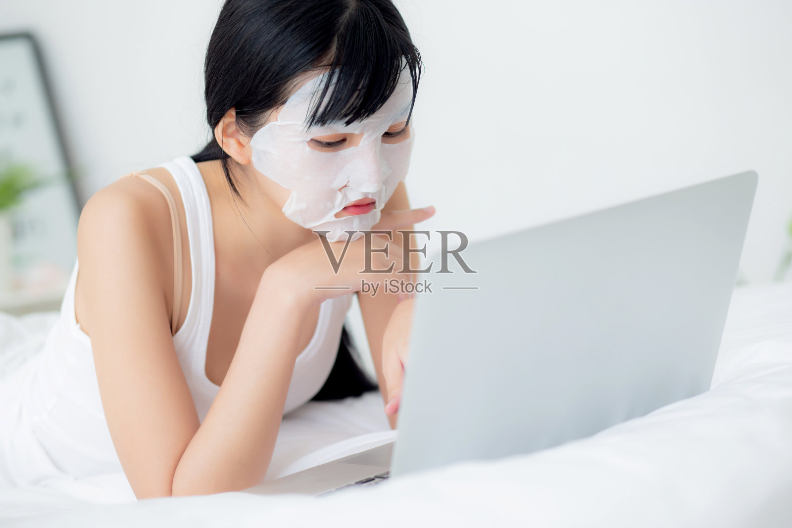 美丽年轻的亚洲女性与床单面膜躺在笔记本电脑上工作在床上的卧室，美容女孩应用面膜的皮肤皱纹在家里，皮肤护理，健康和健康。照片摄影图片