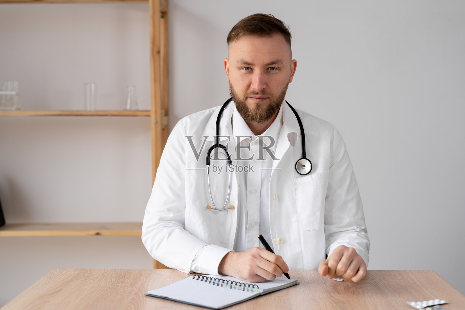 快乐的年轻男医生穿着白大褂，听诊器看着镜头。微笑的医生，全科医生通过视频电话在线咨询病人。照片摄影图片