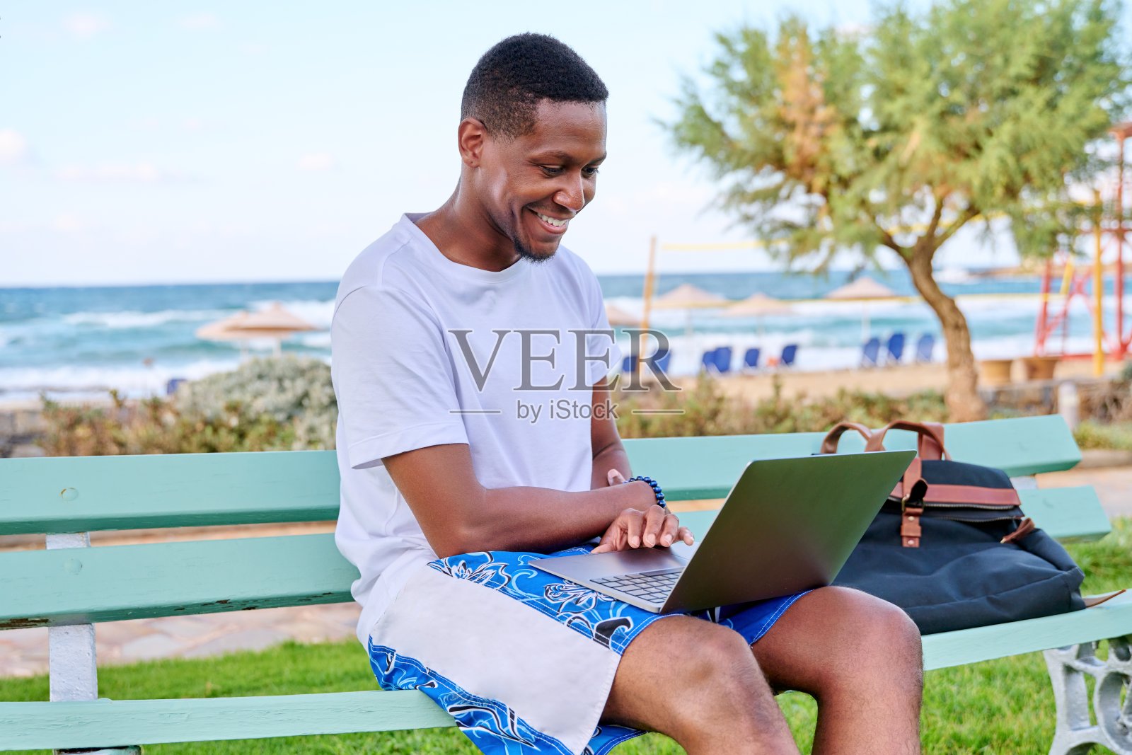 年轻微笑的自由职业者男性在沙滩上放松，坐在长凳上使用笔记本电脑照片摄影图片