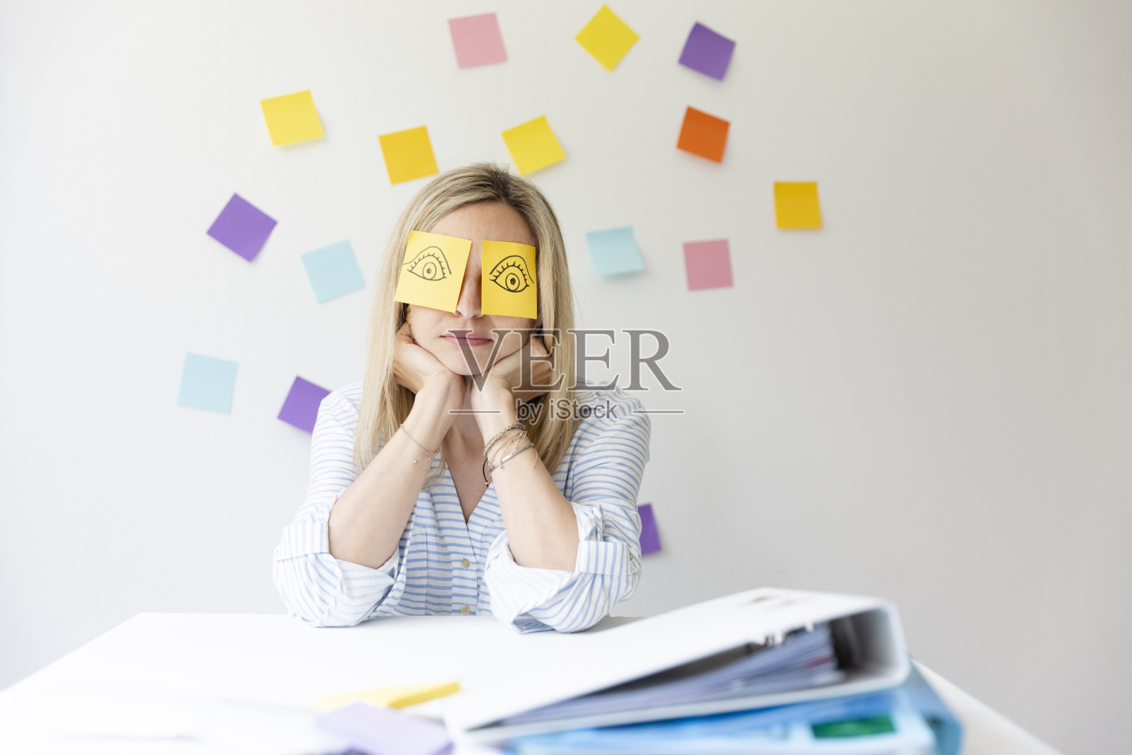 商务女性坐在明亮的现代办公室后面，她的办公桌上有许多办公室的东西，她的脸上有画着眼睛的便签和睡觉照片摄影图片