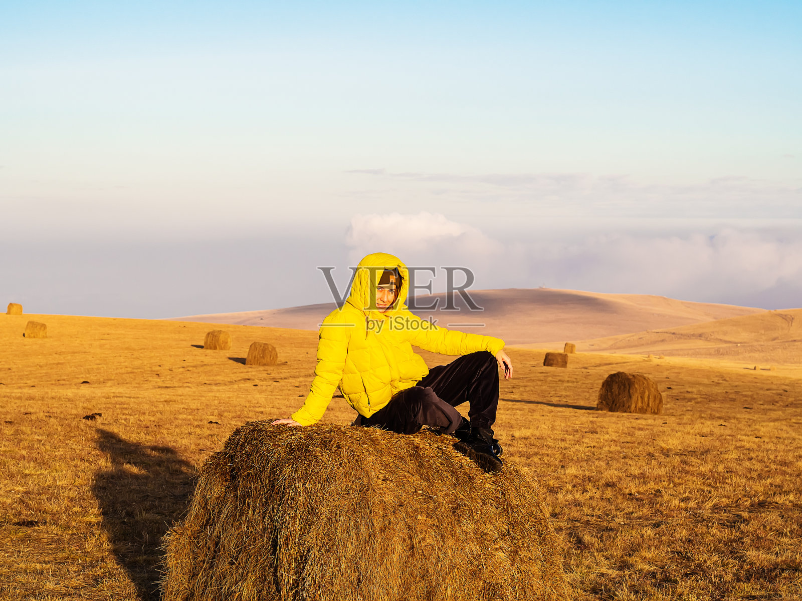 一个女人穿着明亮的黄色温暖的夹克，坐在秋天的田野里，在明亮的阳光照耀下的干草堆上照片摄影图片