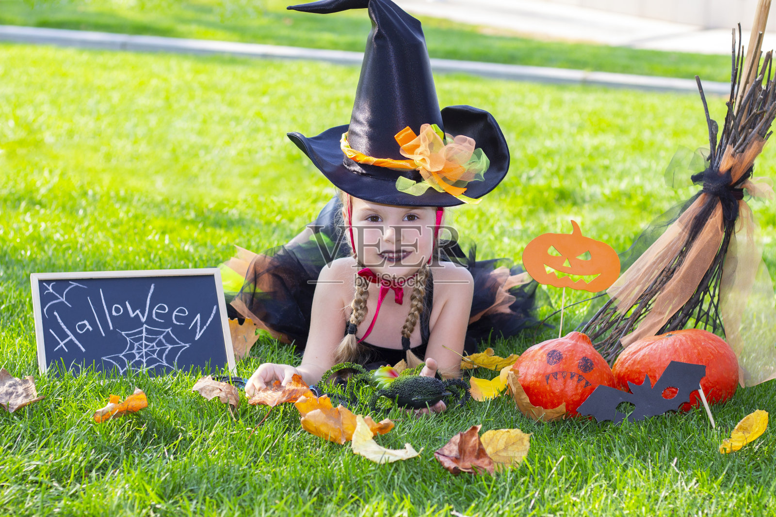 万圣节穿着女巫服装的女孩。贴着标语:万圣节。一个戴着女巫帽的女孩躺在草地上照片摄影图片