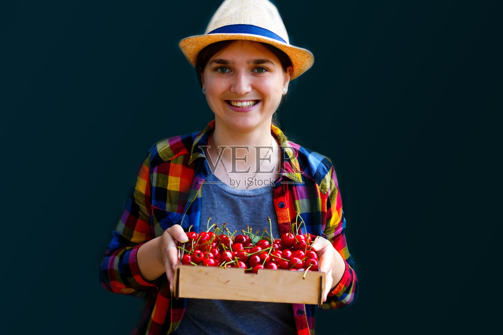 一个心不在焉的年轻女人，戴着帽子，穿着衬衫，微笑着，展示了一篮樱桃和甜樱桃。深绿色背景。夏天的食物,水果。夏天的背景。的焦点照片摄影图片