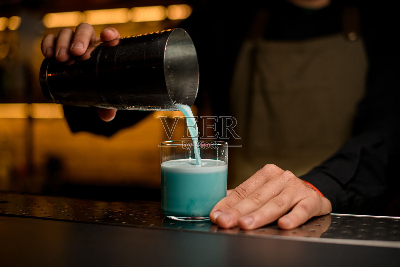 调酒师的男性手从调酒壶中倒出蓝色鸡尾酒的玻璃杯的正面视图照片摄影图片