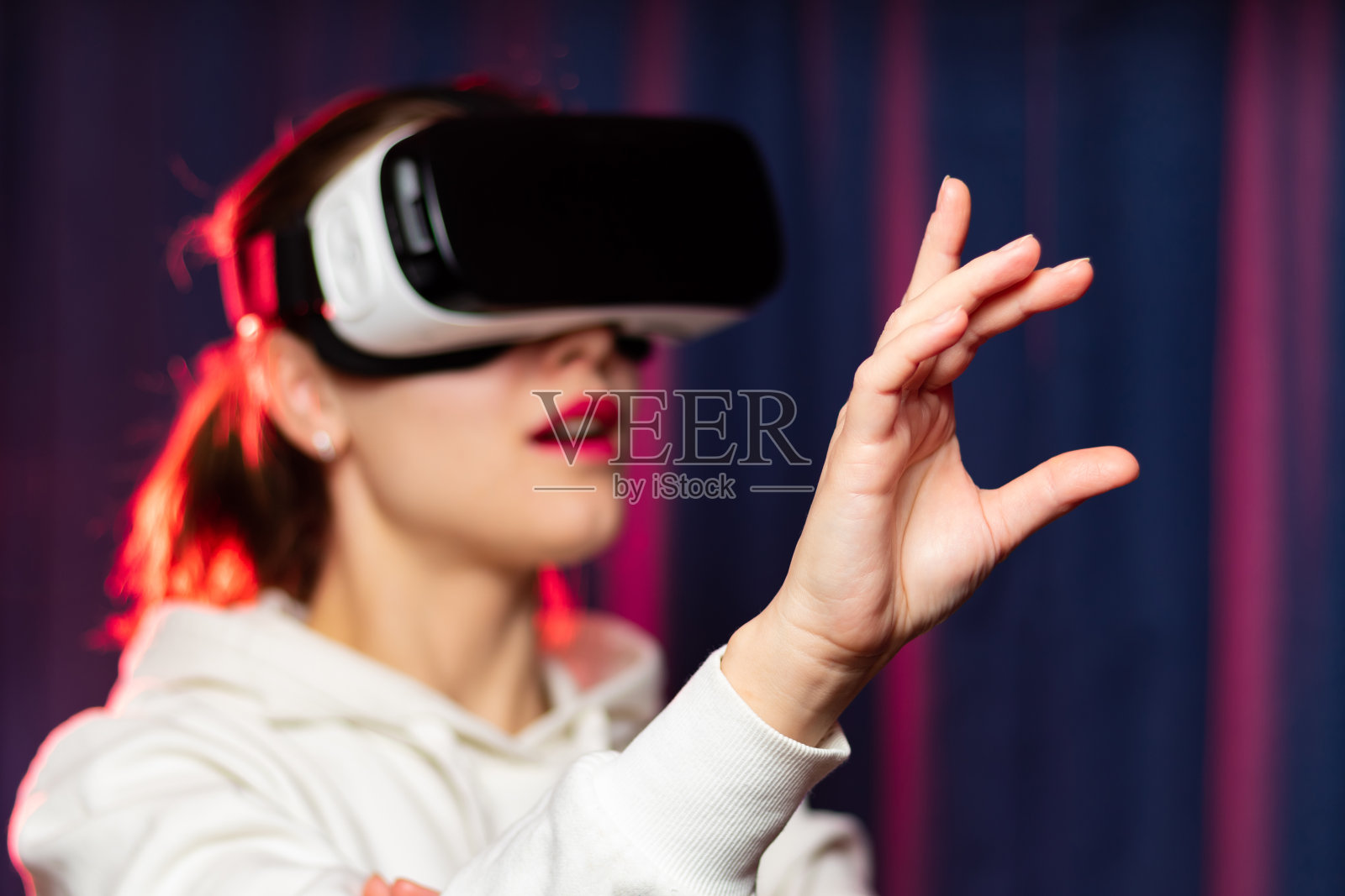 美丽的年轻女子戴着虚拟现实眼镜和白色帽衫在紫色的背景。现代技术。有选择性的重点。特写镜头照片摄影图片