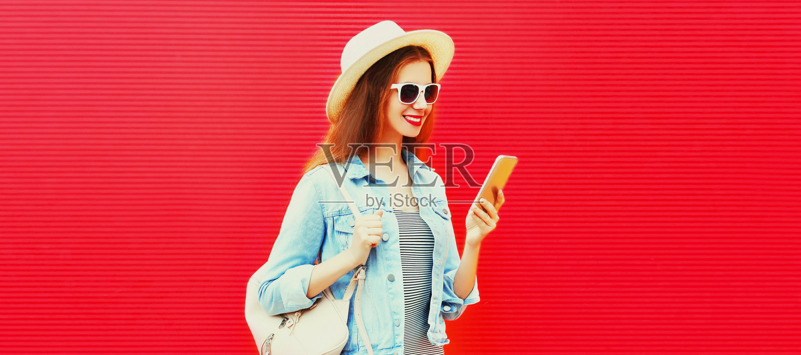 快乐微笑的年轻女性游客的肖像与智能手机戴夏天草帽，背包在红色的背景照片摄影图片