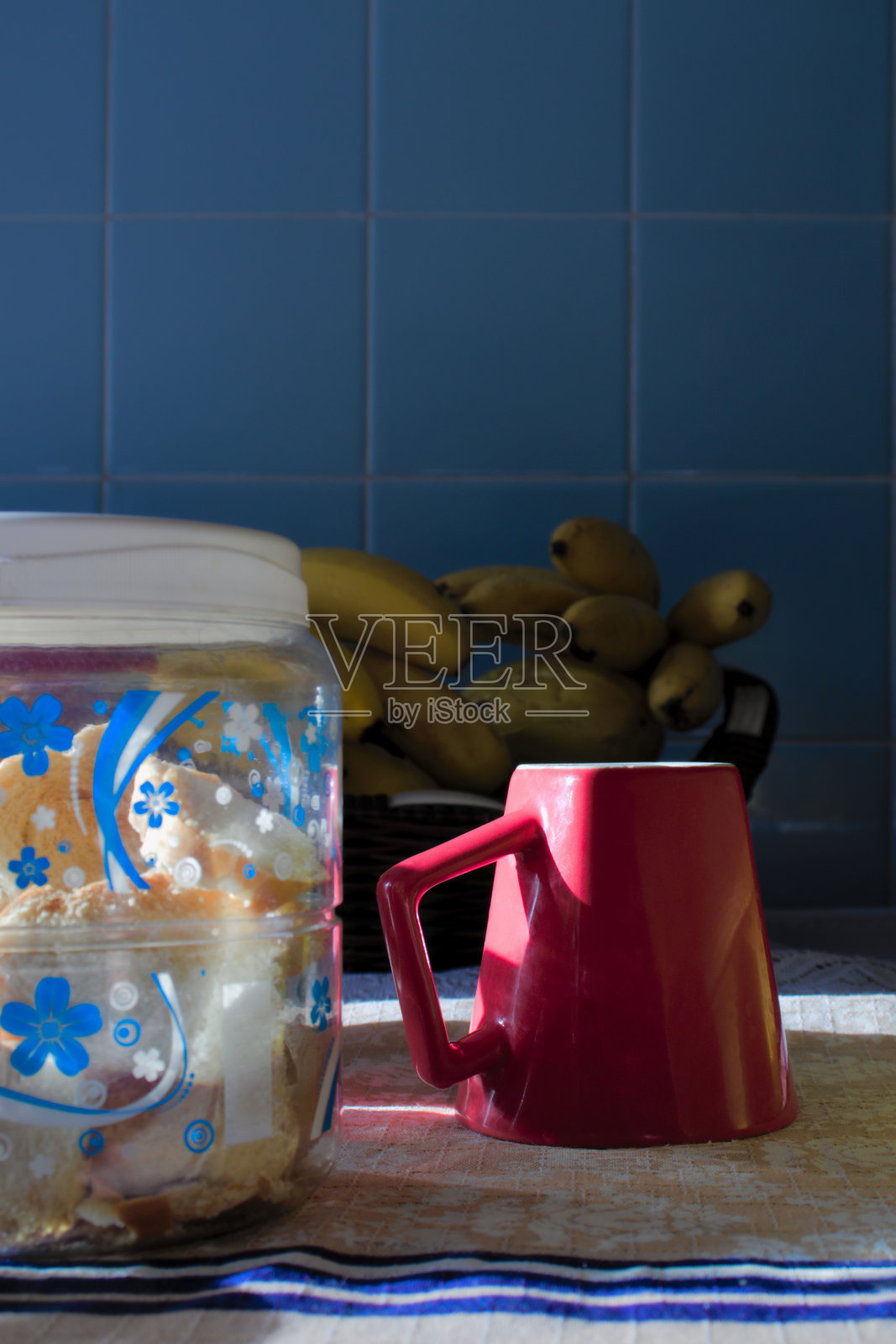 在晨曦的照耀下，桌布上放着红色的陶瓷杯和塑料壶，上面放着饼干。背景是一个模糊的水果盘和一面墙的蓝色瓷砖。照片摄影图片
