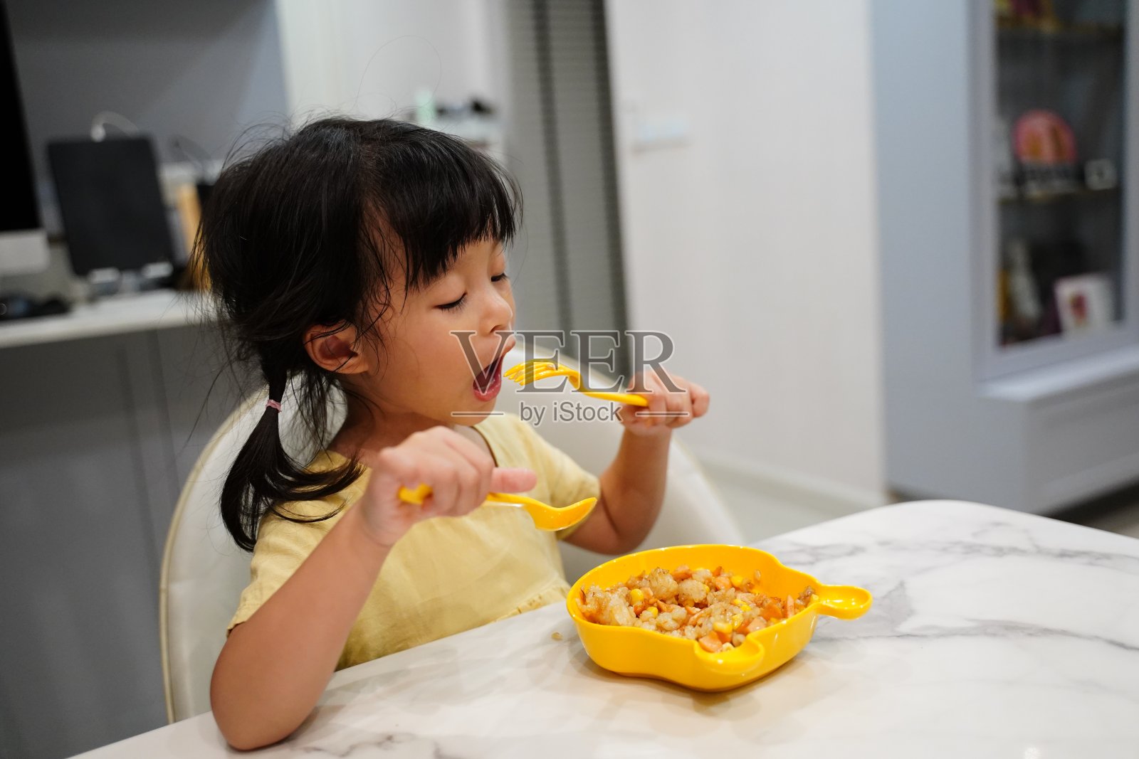 亚洲小女孩自己吃自己坐在餐桌上吃自己的炒饭用黄色的衬衫和黄色的盘子一套勺子和叉子。照片摄影图片