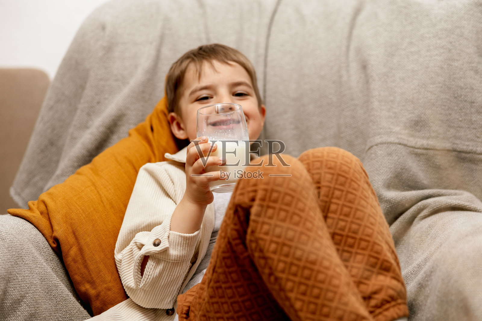 可爱的小男孩坐在家里的沙发上喝牛奶。玻璃鲜奶，乳制品健康饮料。保健，钙，乳糖的来源。学龄前儿童的休闲服装。照片摄影图片