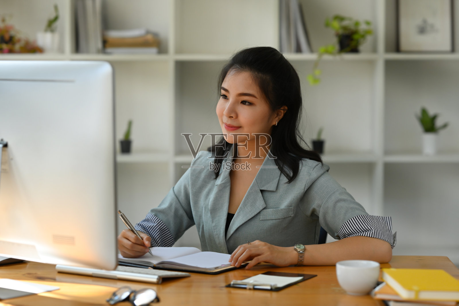 一个漂亮的亚洲女人的肖像坐在办公室里看着电脑和工作笔记，为商业和技术概念。照片摄影图片