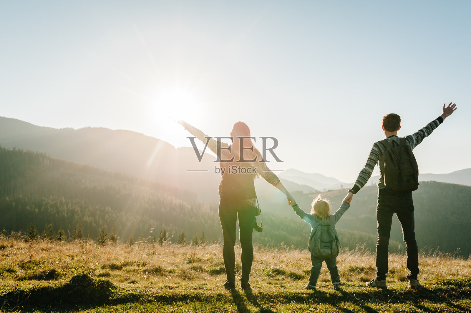 一家人带着孩子爬山。年轻的游客们在山顶上欣赏山谷日落。妈妈开心，爸爸背着双肩包和女儿开心。旅行的概念。世界旅游日。后视图。照片摄影图片