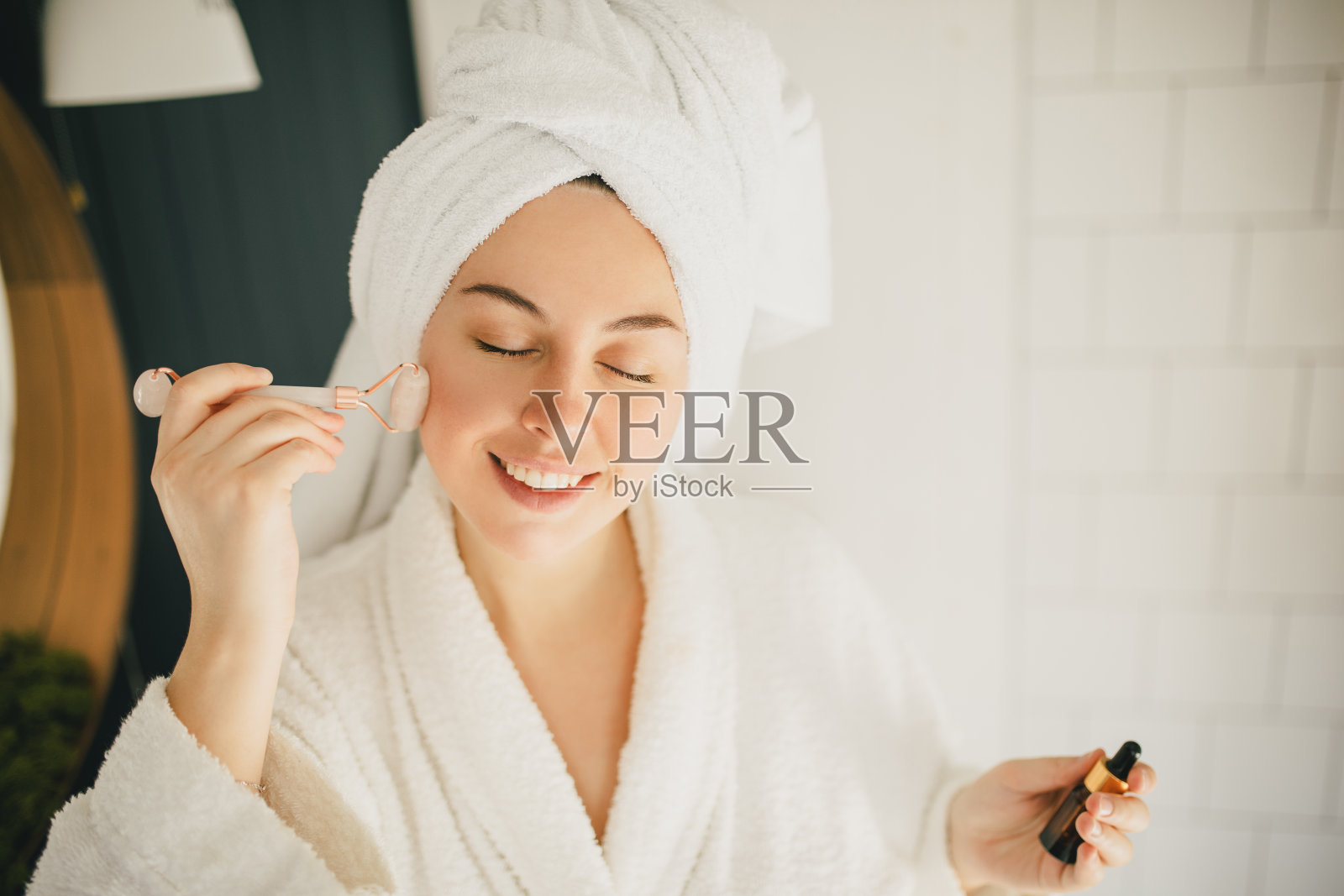 美丽的年轻女子，完美的皮肤，穿着白色浴袍和毛巾在头上淋浴后，在浴室用玉辊做脸部按摩。照片摄影图片