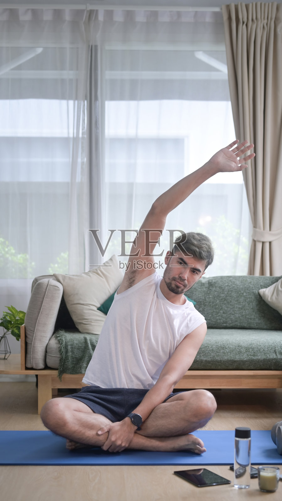 穿着运动服的帅哥坐在家里的瑜伽垫上做热身和伸展运动。照片摄影图片