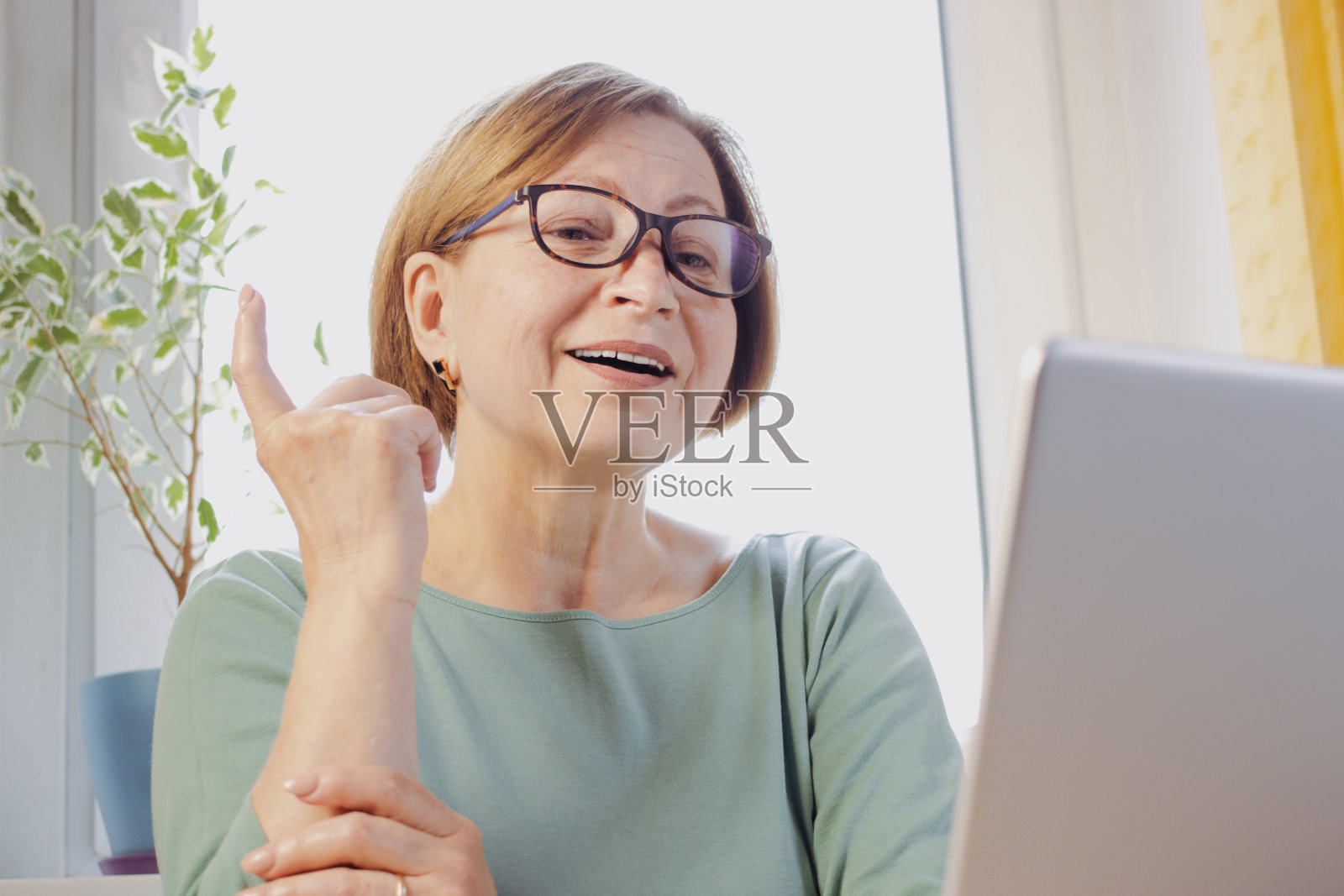 中年快乐的女人坐在桌子前，在笔记本电脑上工作，举起食指，她知道如何解决一个问题，有一个绝妙的想法。老年一代和科技。创造性的工作方法照片摄影图片