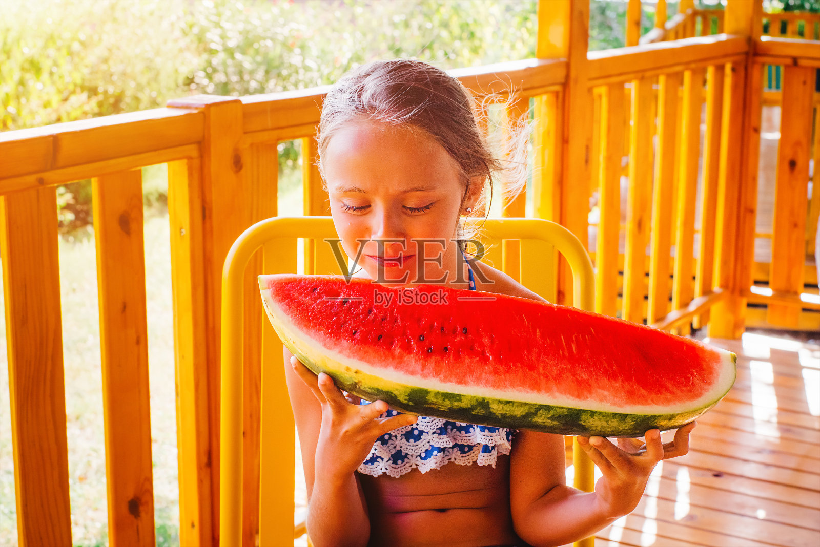 夏日露台上的女孩笑着吃了一大块红色的西瓜。自然的食物。女孩在夏天吃一个成熟多汁的西瓜。可爱的金发女孩在户外吃一片西瓜。照片摄影图片
