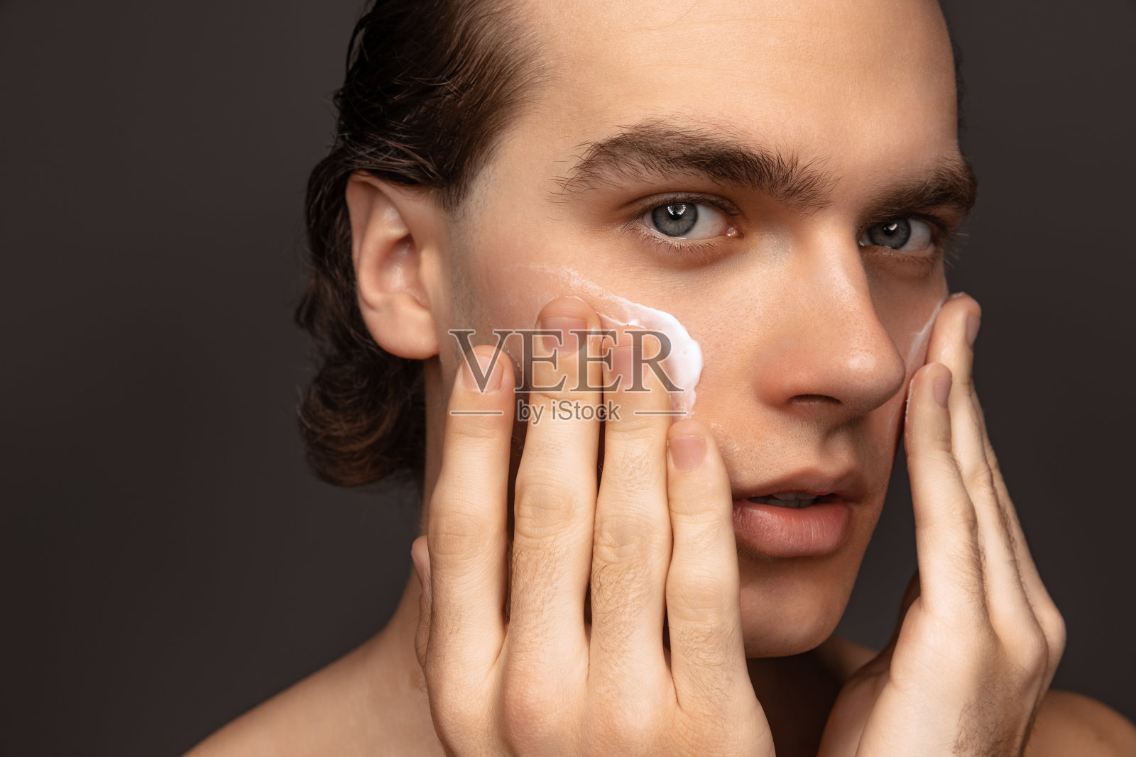 男士面部皮肤护理。年轻快乐的人与完美的保持皮肤使用面霜孤立在灰色的背景。化妆品、保健、护肤、美容照片摄影图片