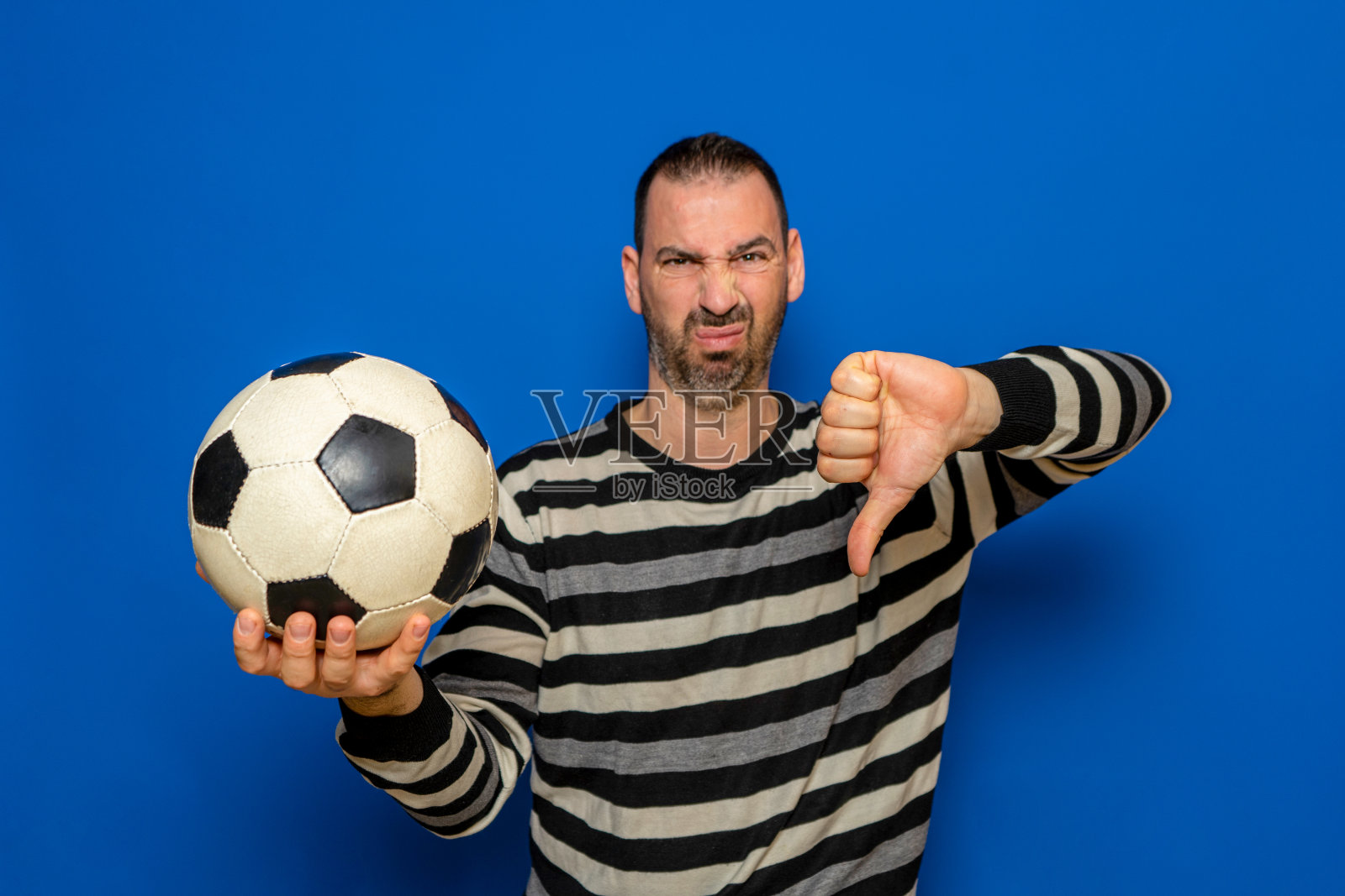 英俊的拉丁男子拿着足球与愤怒的脸，消极的标志显示厌恶与拇指向下，拒绝概念。孤立在蓝色背景。照片摄影图片