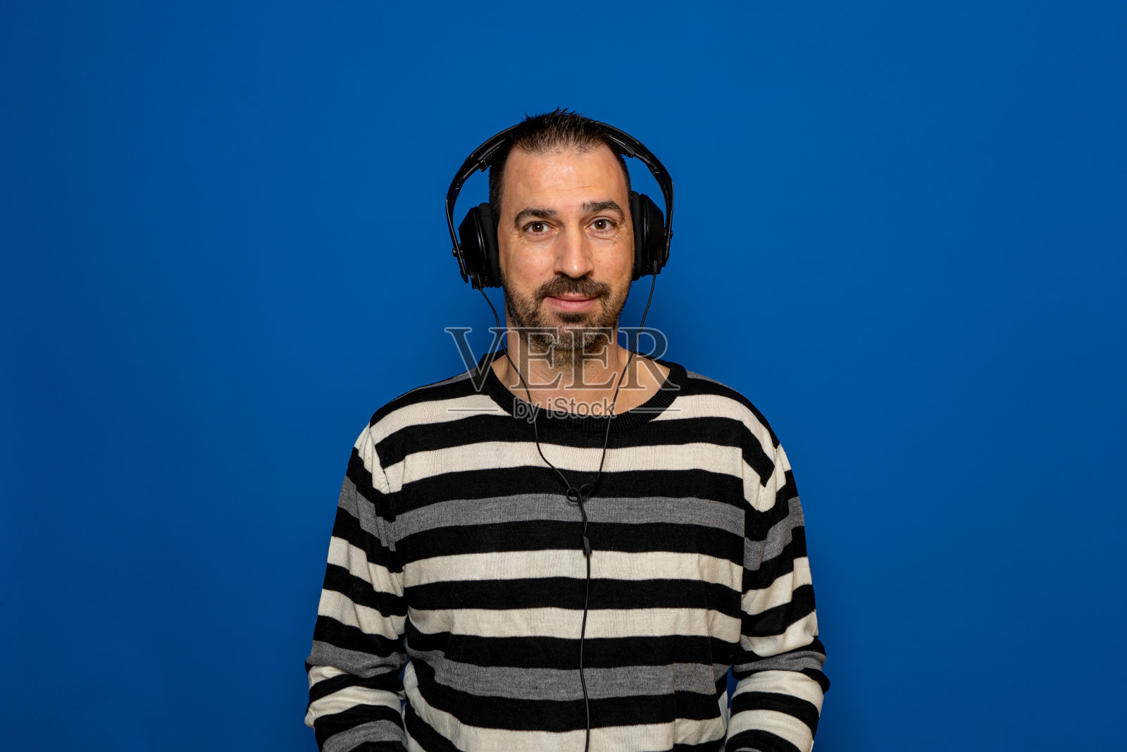 一名留着胡子的西班牙男子穿着一件条纹毛衣，戴着耳机听音乐，在蓝色的录音室背景上隔离照片摄影图片