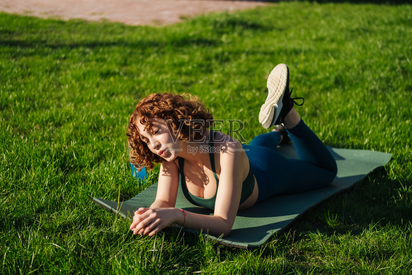 微笑的年轻红发女子拿着手机，在锻炼后放松。女性健身女孩看着相机躺在垫子上。体育，技术和健康的生活方式的概念。照片摄影图片