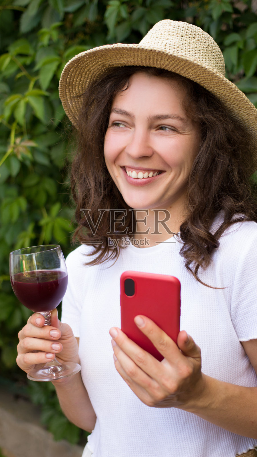 年轻的黑发女人的肖像在白色t恤和夏天的帽子，微笑着举杯，拿着红酒和智能手机，笑着看远处站在绿色的花园。垂直照片摄影图片