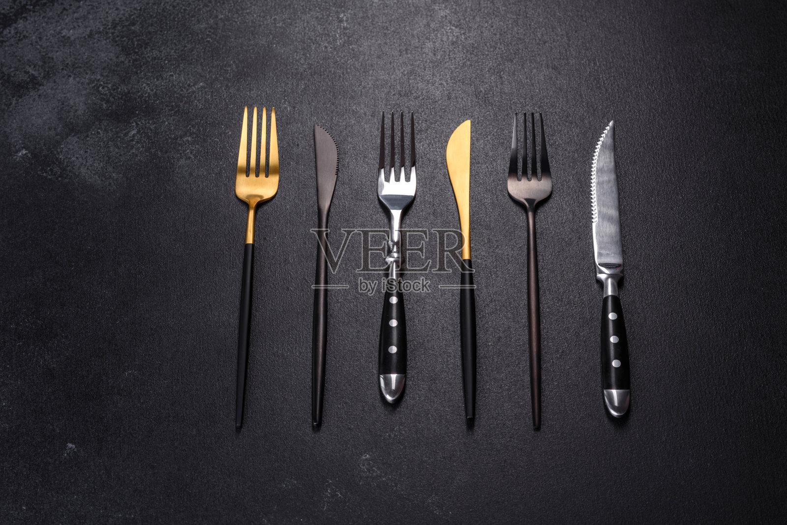 黑色的混凝土背景上摆放着几把黑色、金色和银色的刀叉照片摄影图片
