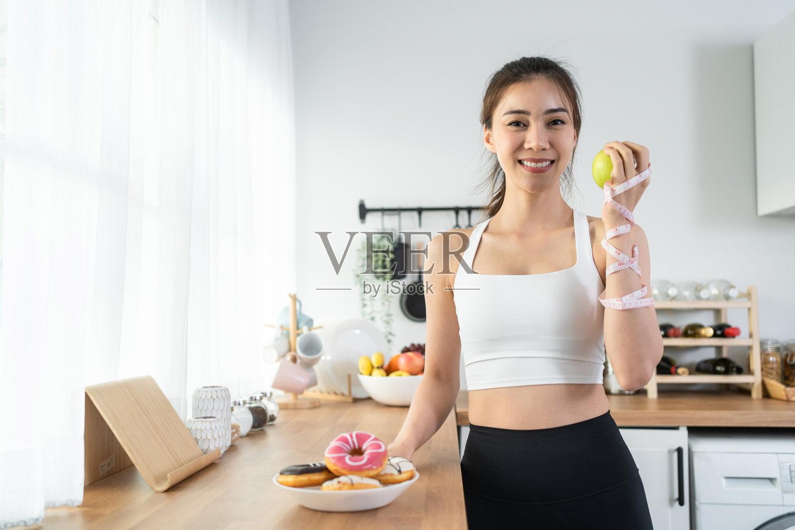 亚洲有吸引力的活跃的女人拿着红苹果在家里的厨房。活跃的运动女孩的肖像在运动服享受吃水果，健康的食物，为健康锻炼后，看相机在房子里。照片摄影图片