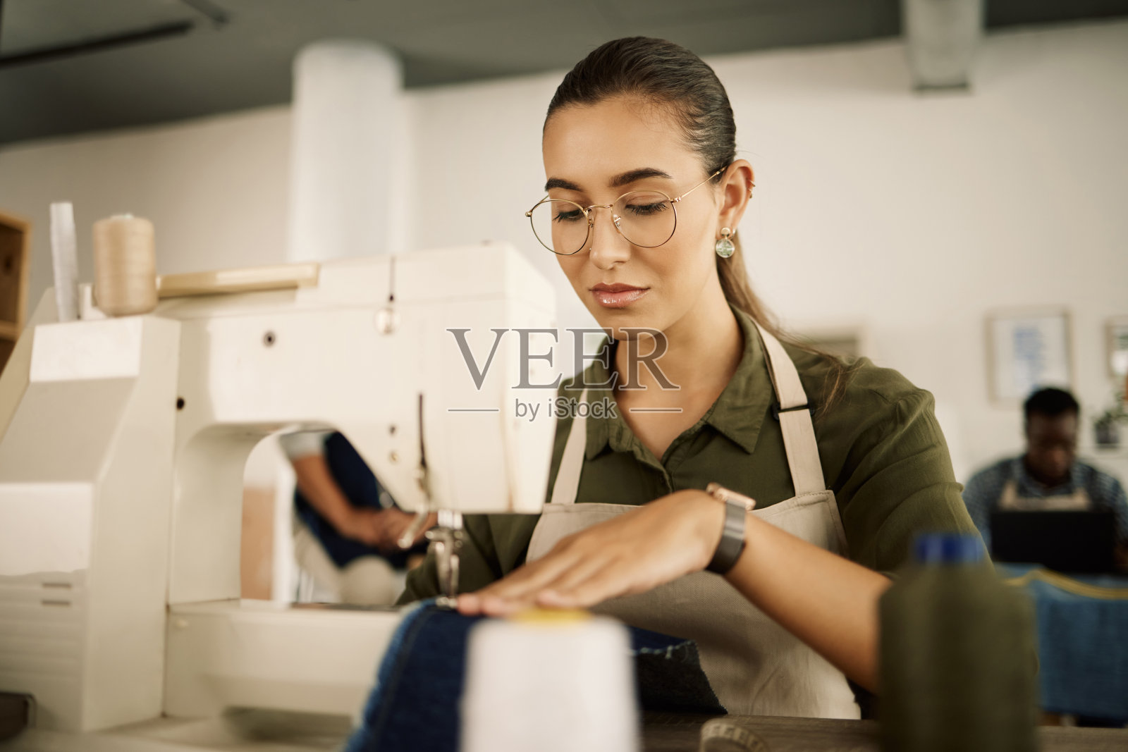 混血设计师在机器上缝纫。年轻时装设计师缝制牛仔面料。在她工作室里工作的严肃裁缝。在缝纫机上缝制材料的创意企业家照片摄影图片